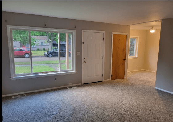 A room with a carpeted floor, a white door, a window with a view of a street, and a wooden door.